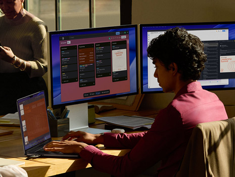 Des collègues assis autour d’une table dans un bureau. Une femme assise à son bureau travaille avec un MacBook et plusieurs écrans. Ses collègues discutent autour d’elle.