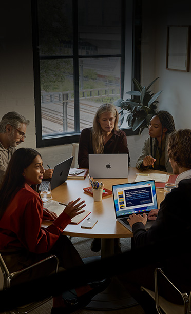 Five individuals seated around a conference table, collaborating on MacBook laptops, with iPhone devices face down on the table