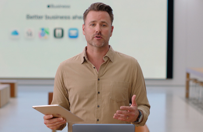 Un presentador habla mostrando un iPad, y al fondo se muestra una presentación con varios íconos de apps para empresas en una pantalla grande.