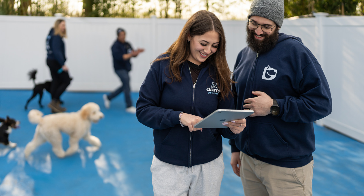 Dos personas con sudaderas de la empresa miran un iPad juntas en un área de juego para perros, con perros y personal en el fondo