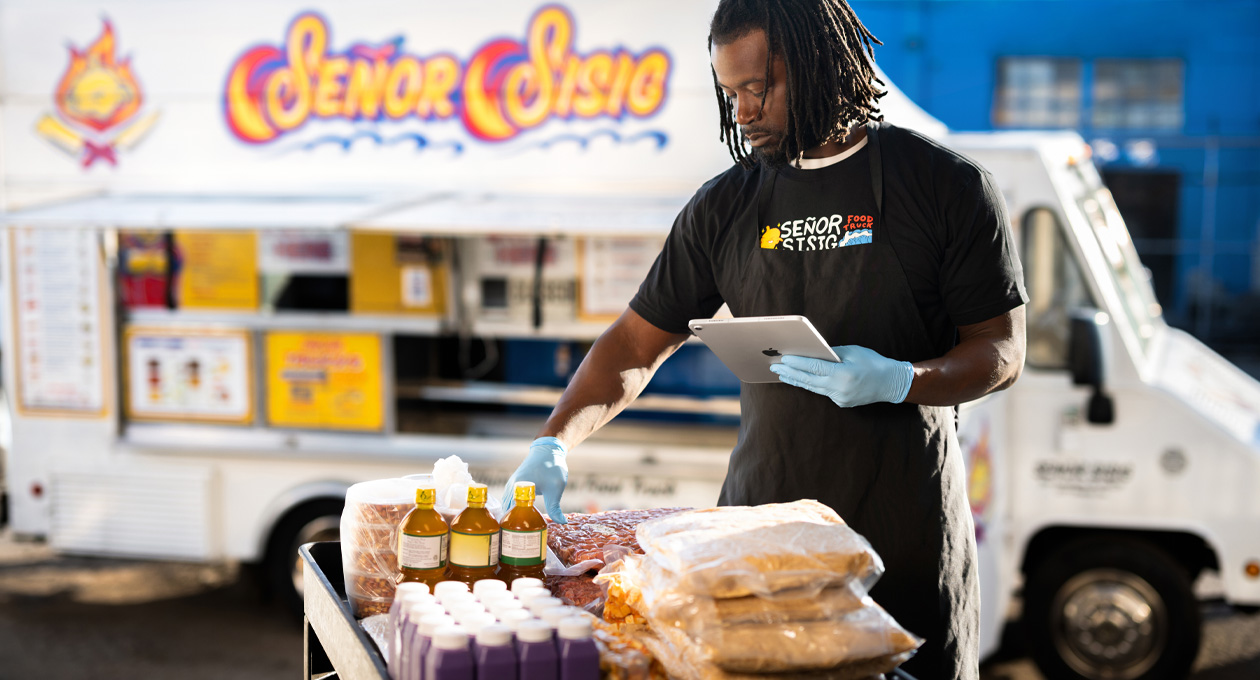 Una persona con una camiseta de la empresa y guantes revisa un iPad mientras prepara comida en una mesa, con una food truck de fondo