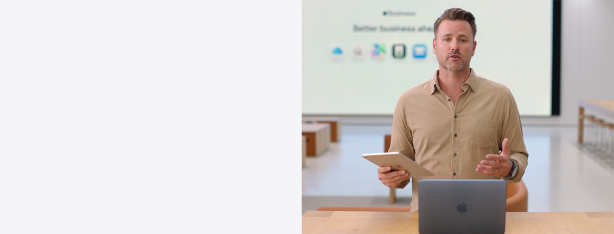 Presenter speaks while holding an iPad and standing in front of a MacBook on a table, while a large screen displays various business app icons.