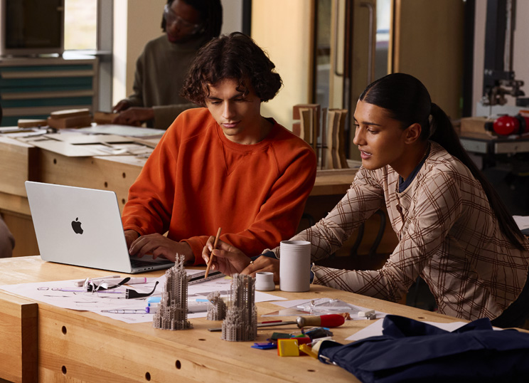 Två personer samarbetar på ett projekt vid ett bord, en skriver på en MacBook, den andra använder penna och linjal på papper, omgivna av konstnärsmaterial.