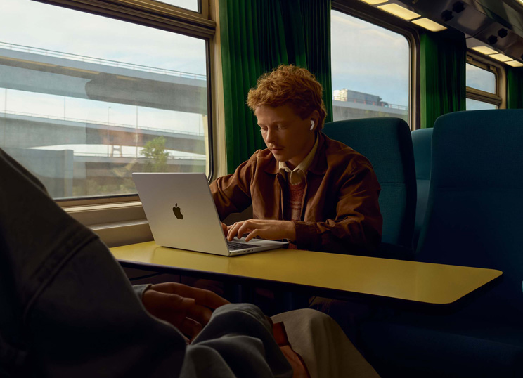 Person på ett tåg vid en fönsterplats som använder AirPods och arbetar på en MacBook.