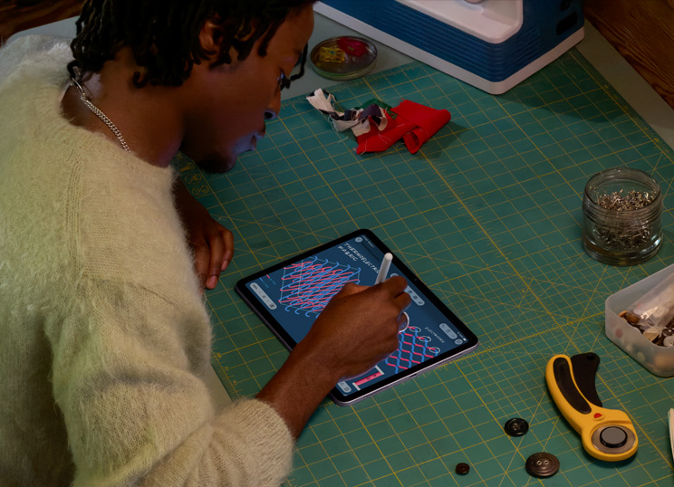 En person som illustrerar med Apple Pencil på iPad, sittande vid en grön skärmatta med symaterial.