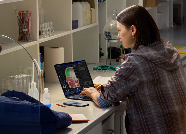 A person sitting in a lab using MacBook, screen displaying multiple apps.
