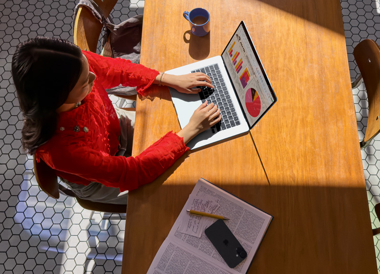A person using MacBook at a table, with a mug, smartphone, and papers nearby.
