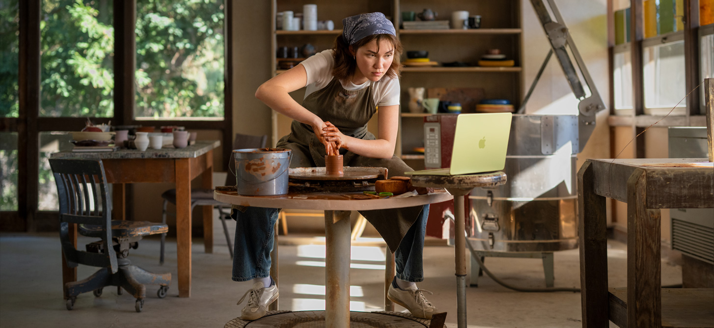 An art student shapes clay at a pottery wheel referring to an open MacBook Neo, highlighting durable design and ability to work anywhere