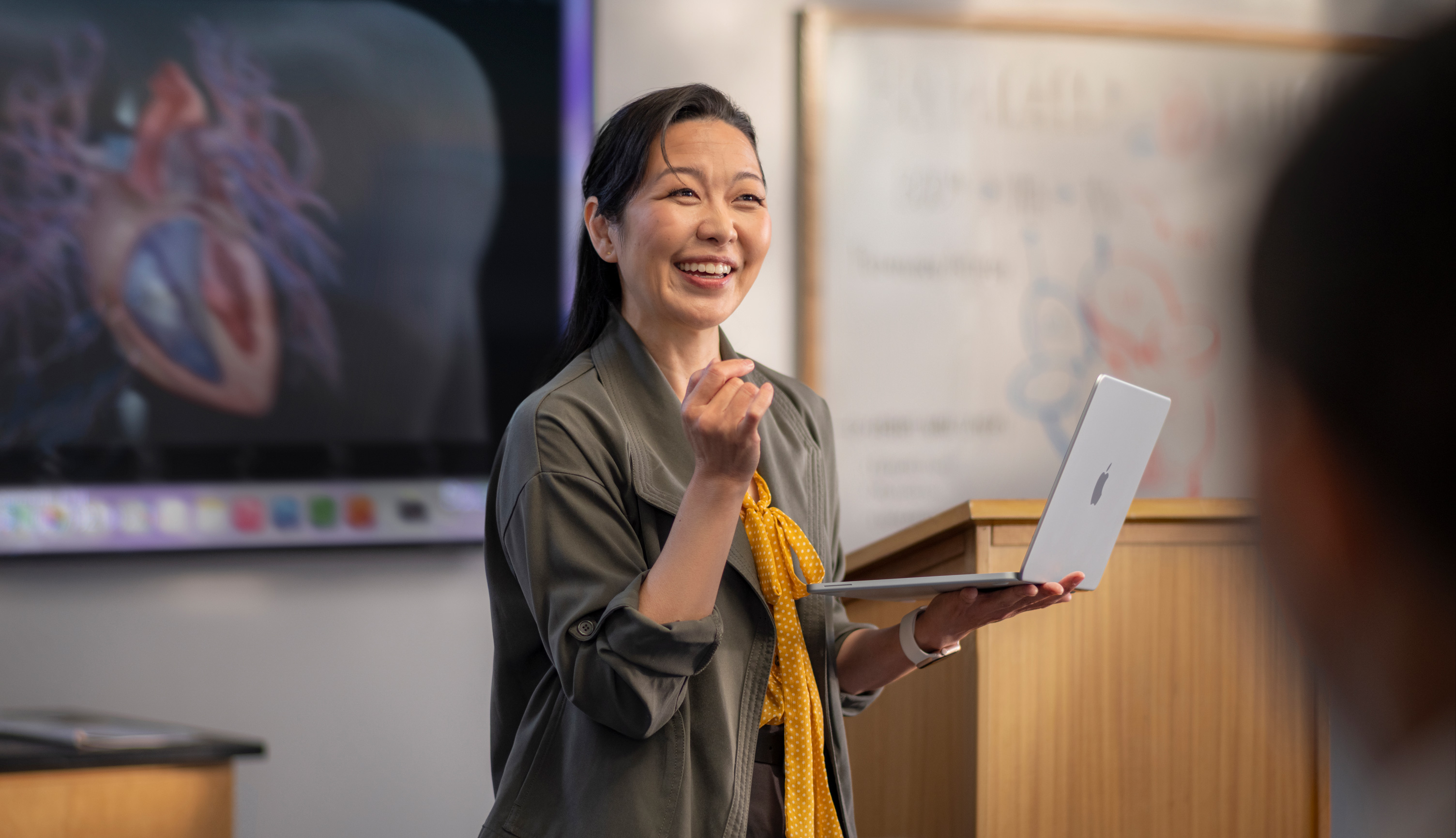 A professor gives a presentation in a lecture hall while holding a MacBook, demonstrating performance and long battery life