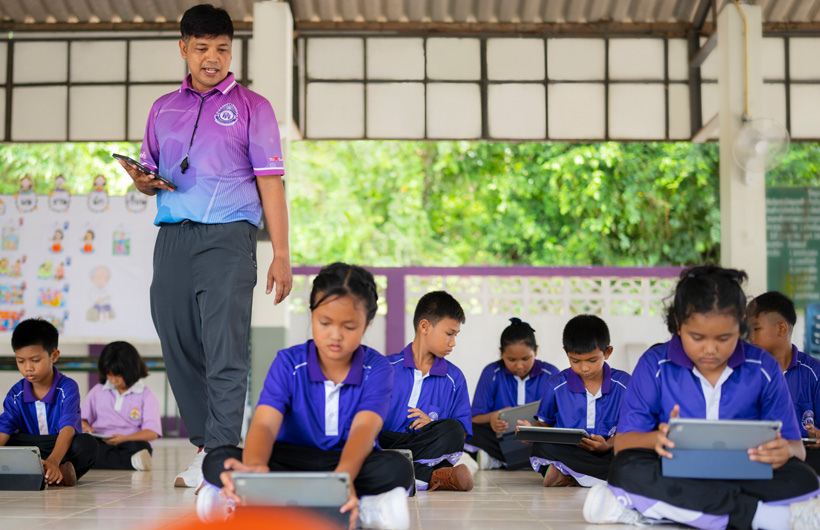In an outdoor classroom, a physical education teacher walks among his students, who are seated on the ground using iPad devices.