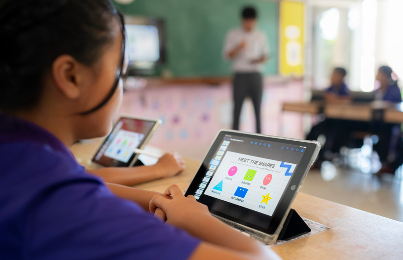 Close‑up of a student at their desk, using an iPad. On screen, there are colourful shapes displayed with the title, “Meet the shapes”.