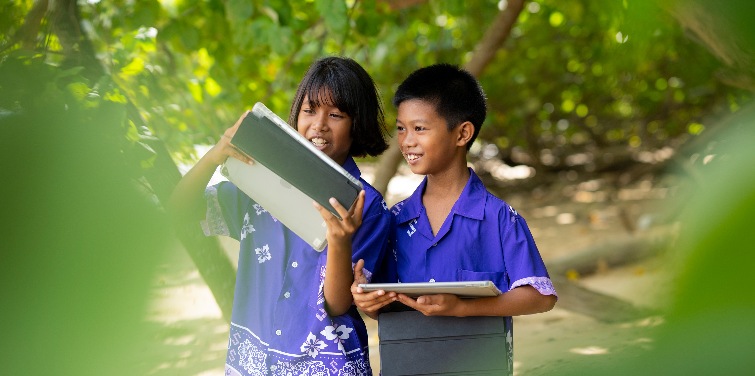 Two primary students, one boy and one girl, standing outside amidst tropical trees, each holding an iPad. They are both looking at the girl’s iPad screen, smiling and engaged.