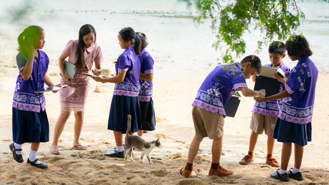 Six primary students in colourful purple uniforms stand on a sandy beach holding iPad devices. A teacher is assisting one student on an iPad, and there is a cat walking amongst them.