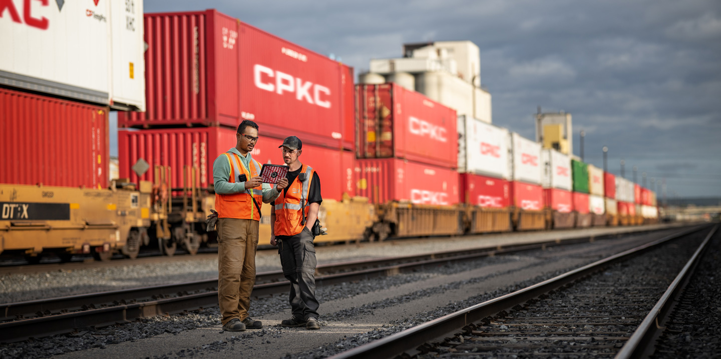 Two railroad workers reference an iPad while standing in a train depot beside a stationary train carrying shipping containers