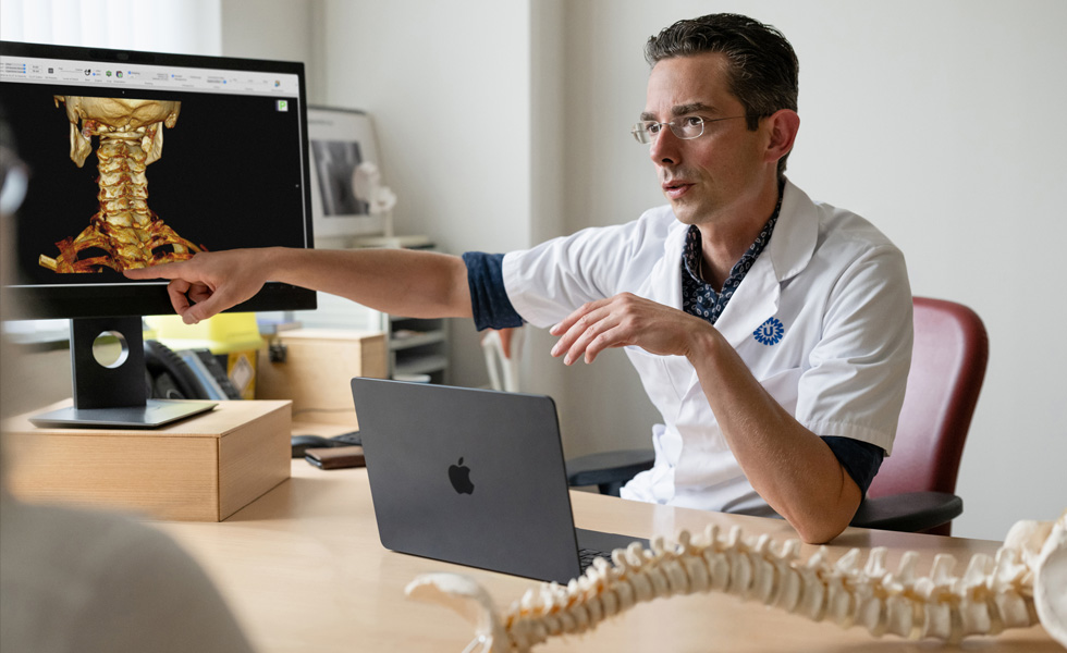 Un professionnel de santé montre un rendu 3D des os de la nuque sur un Studio Display, tandis qu’un MacBook est posé sur son bureau