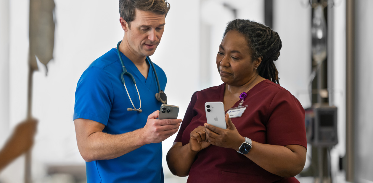 Een mannelijke zorgverlener in blauwe kleding laat iets op zijn iPhone zien aan een vrouwelijke zorgverlener in rode kleding, die ook een iPhone heeft.